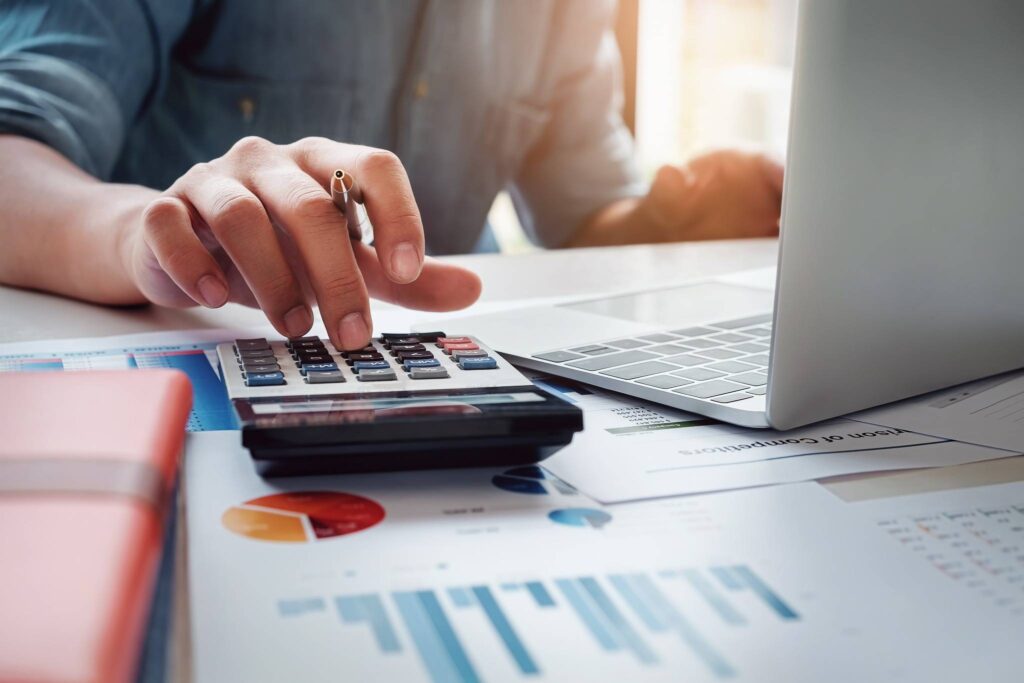 Hand van ondernemer met pen werkt aan exploitatiebegroting met laptop, calculator en financiële overzichten