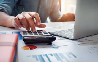 Hand van ondernemer met pen werkt aan exploitatiebegroting met laptop, calculator en financiële overzichten