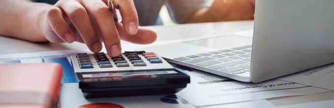 Hand van ondernemer met pen werkt aan exploitatiebegroting met laptop, calculator en financiële overzichten